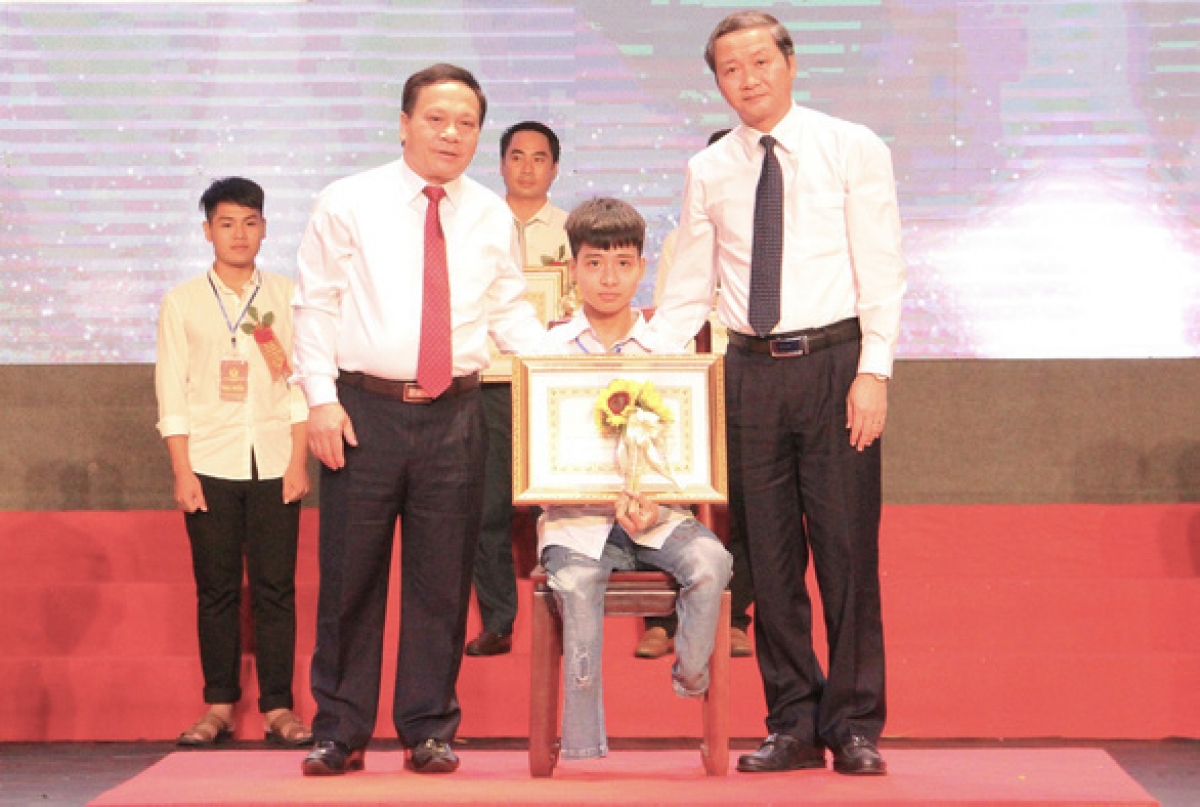 Nguyễn Tất Minh bị khuyết tật ở chân (người ngồi ghế) nhận bằng khen của chủ tịch UBND tỉnh Thanh Hóa tại Đại hội thi đua yêu nước chiều 2-10. Ngô Văn Hiếu (đứng sau), người đã cõng Minh đi học suốt 10 năm, cũng được khen trong đợt này. Ảnh: Hà Đồng.