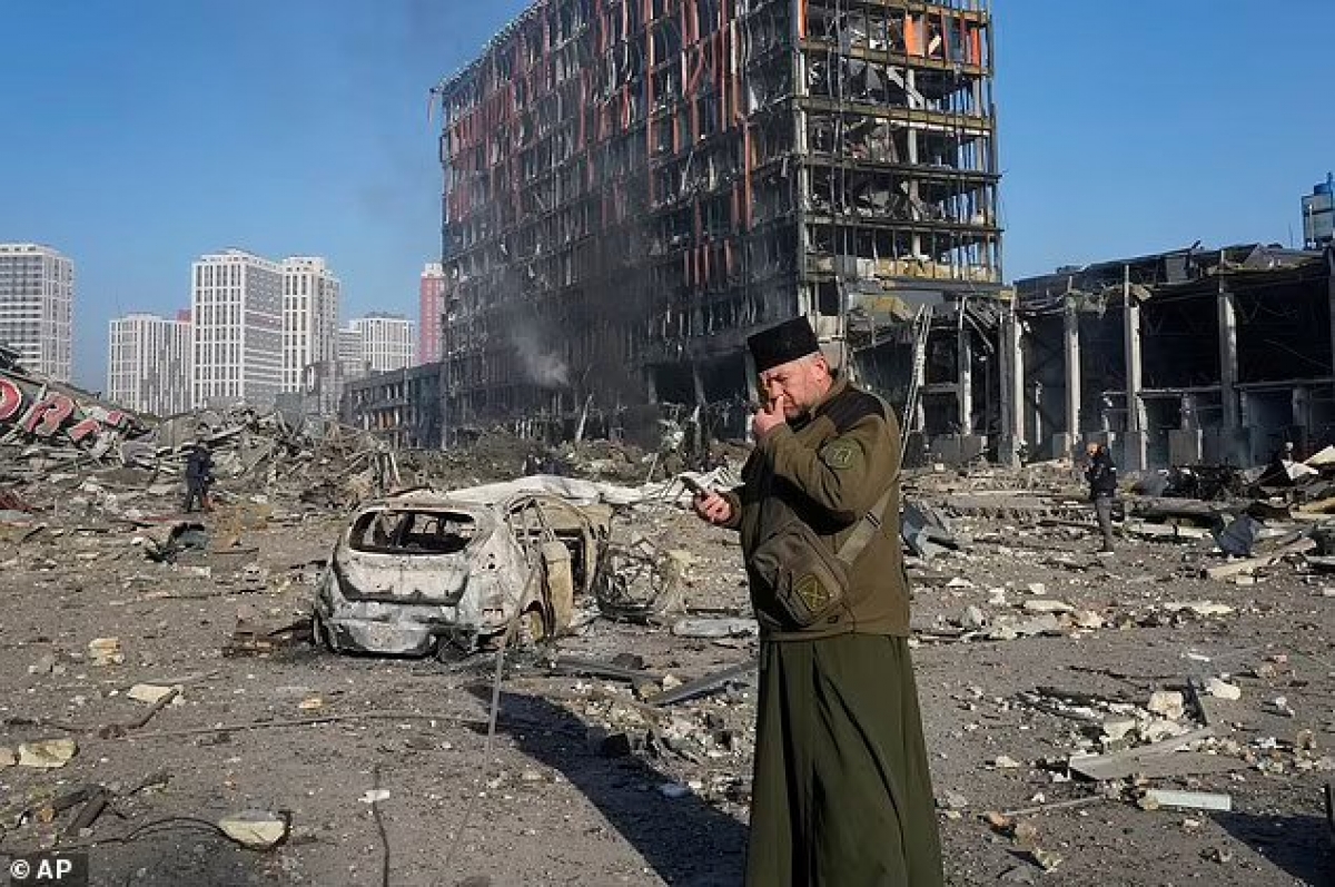 Một linh mục đang cầu nguyện trước trung tâm mua sắm tan hoang vì bom đạn ở Kyiv. (Ảnh: AP).