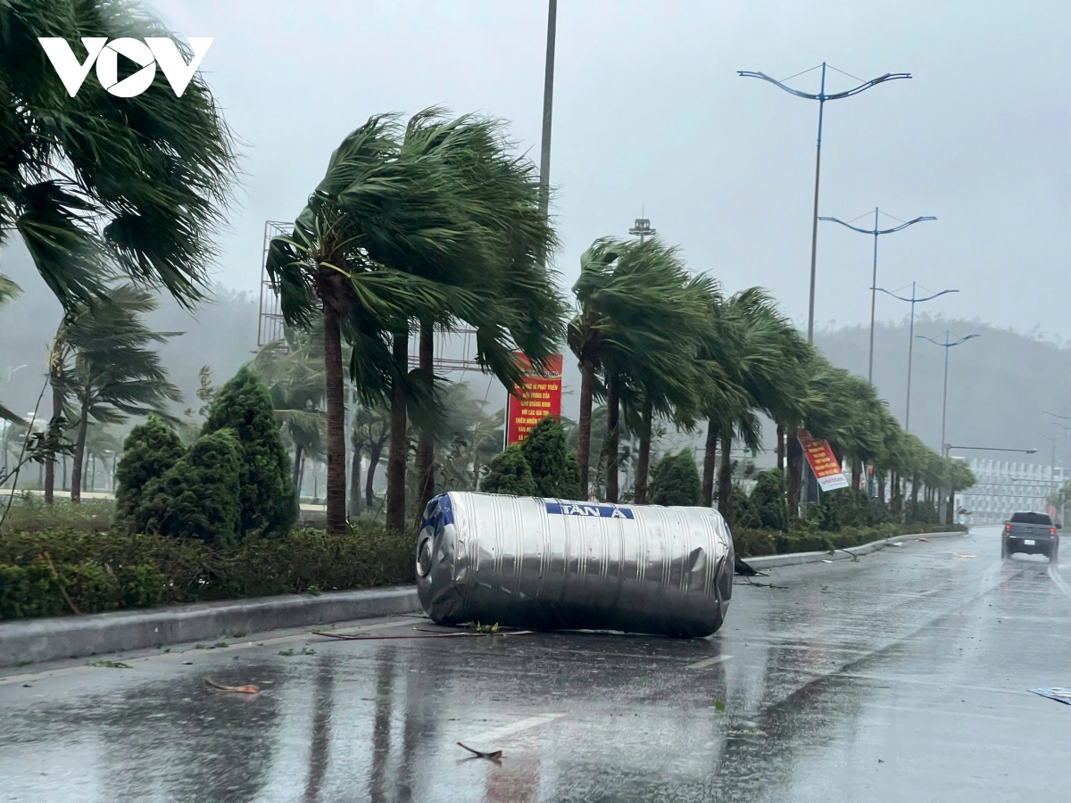 The strong wind blows a large water tank from a building onto the street, creating a hazard for moving vehicles.