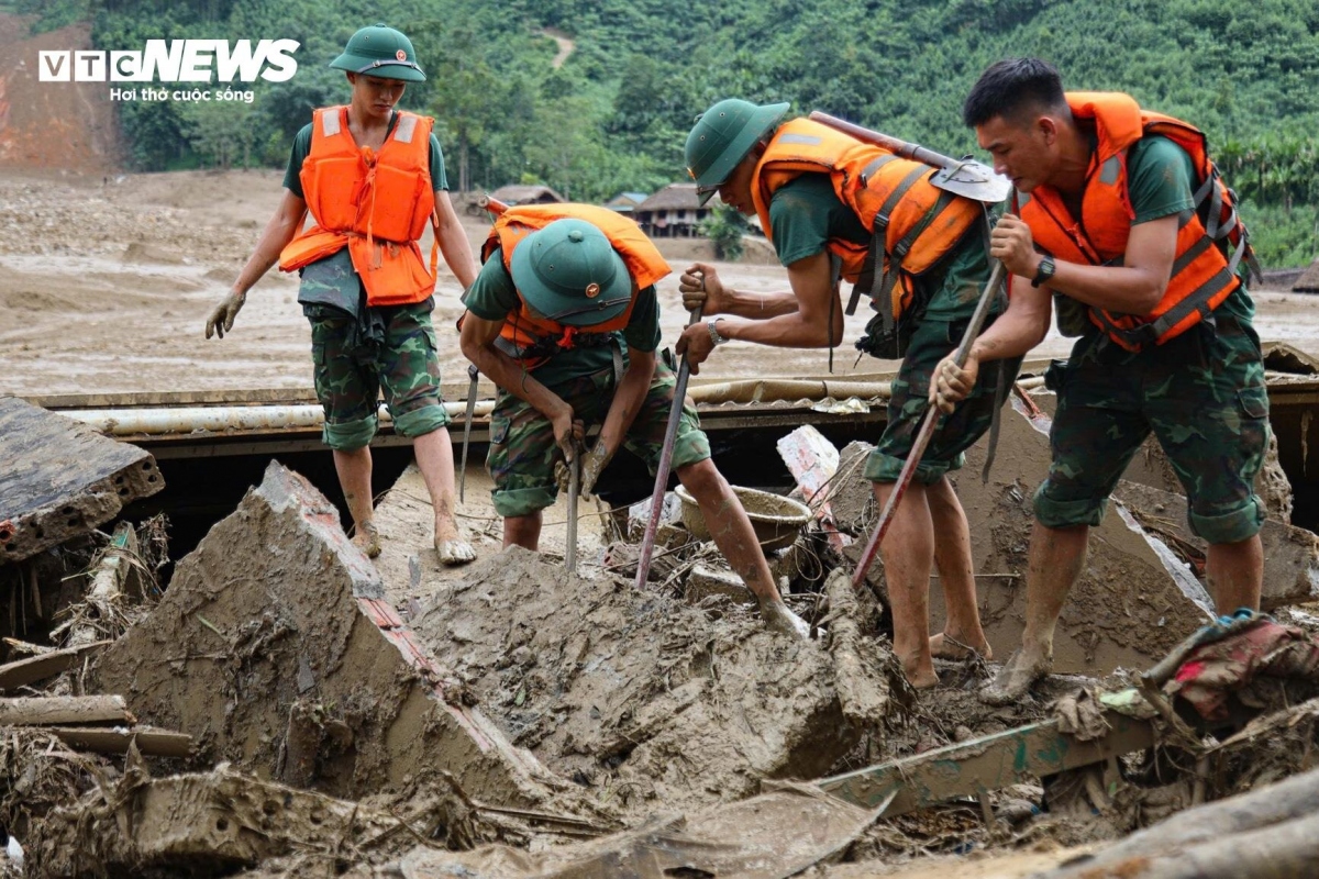 The soldiers carefully navigate through the thick mud, avoiding countless sharp objects like nails, metal pieces, and even everyday knives from houses that have been swept away by the flashfloods.