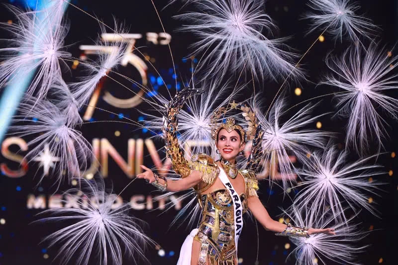 oa hậu Paraguay, Naomi Méndez, đã có màn biến hóa trang phục ấn tượng khi sải bước trên sàn diễn. Người đẹp 32 tuổi này xuất hiện trên sân khấu trong chiếc váy trắng nhiều lớp, đội mũ vàng và đeo một kết cấu tròn trên vai với những cây hoa trắng được gắn kèm. Nhưng chỉ với một vòng xoay, cô đã cởi bỏ lớp trên cùng và để lộ bộ đồ liền thân màu vàng trông giống như một bộ áo giáp.