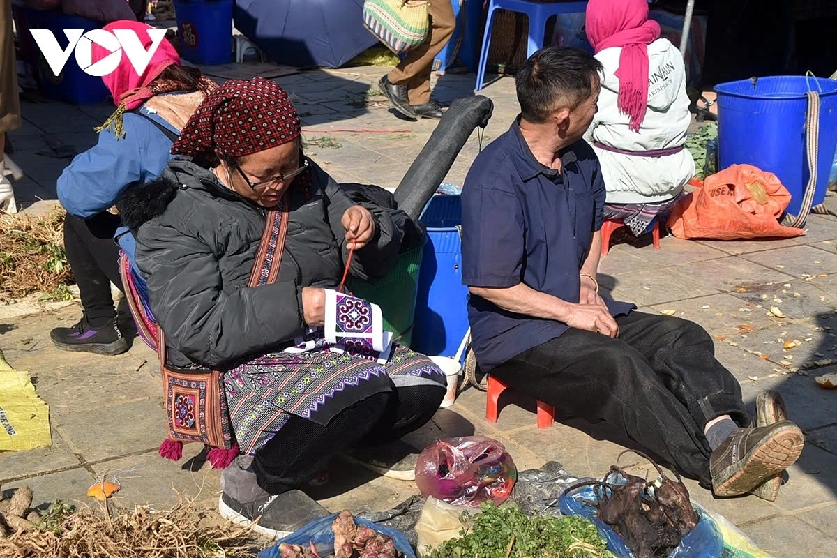 A woman calmly sits in the middle of the busy market, embroidering a colorful traditional dress.