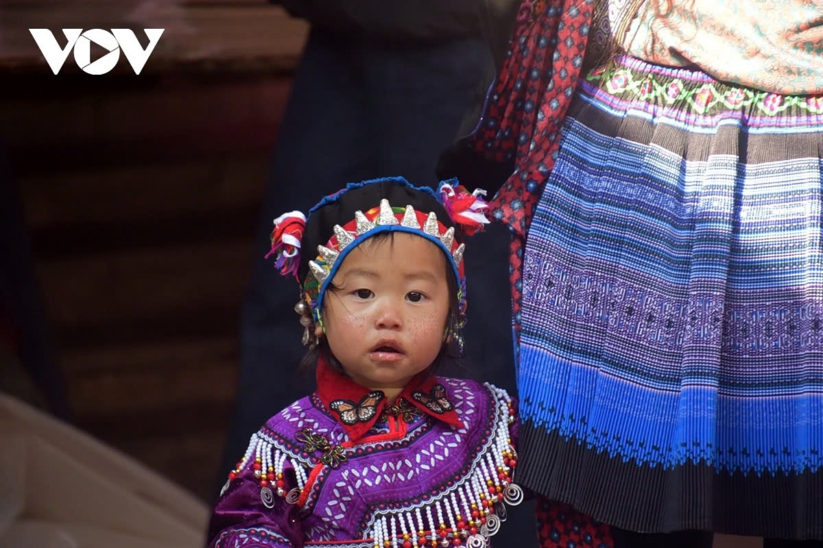 A H'mong child innocently curls up next to her mother’s skirt, watching the vibrant world around her.