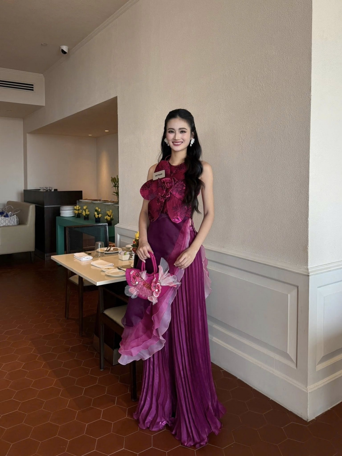 Ở ngày tiếp theo tại đấu trường nhan sắc quốc tế, Hoa hậu Ý Nhi diện một thiết kế khá cầu kỳ, màu tím hồng đậm với bông hoa bản to đính kết ở ngực. Cô kết hợp với chiếc túi cùng màu, được trang trí tỉ mỉ. 
