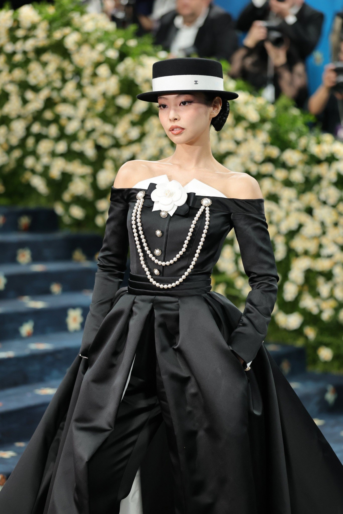 Jennie hóa thân thành quý cô Hollywood cổ điển trong bộ váy tuxedo đen trễ vai từ Chanel, kết hợp hoa trà và chuỗi ngọc trai đặc trưng của nhà mốt Pháp. Điểm nhấn là chiếc mũ boater và kiểu tóc búi thanh lịch, gợi lên hình ảnh giới quý tộc thời thượng.