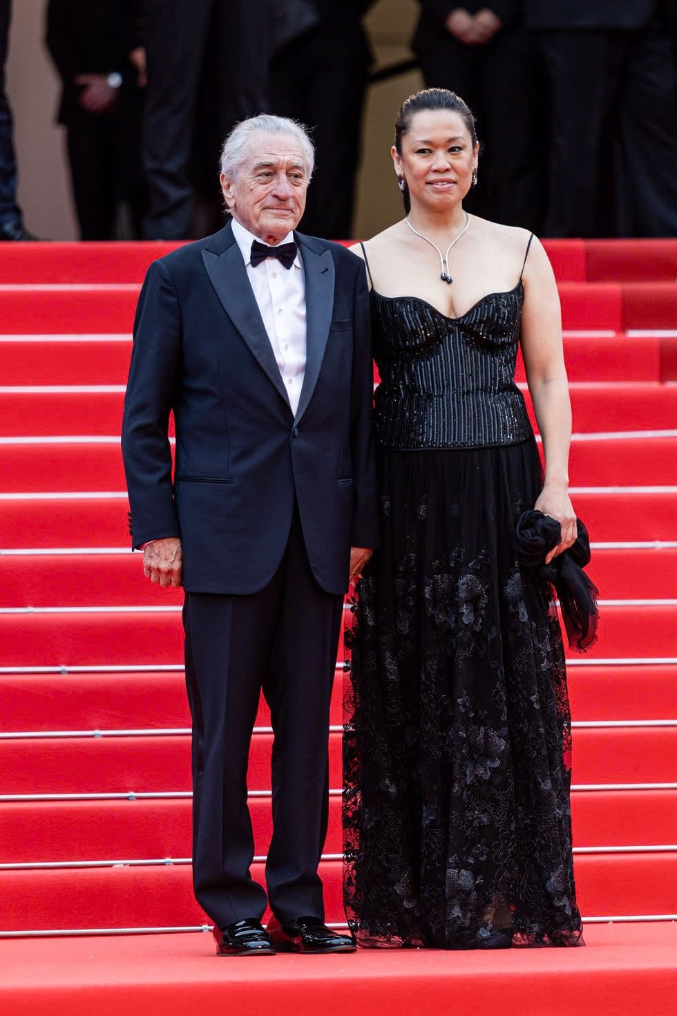 Robert De Niro lịch lãm trong bộ vest xanh đậm phối cùng phụ kiện màu đen và bạn gái kém 36 tuổi Tiffany Chen thanh lịch với chiếc váy đen hai dây, phần thân áo đính sequin lấp lánh và chân váy thêu hoa tinh tế.